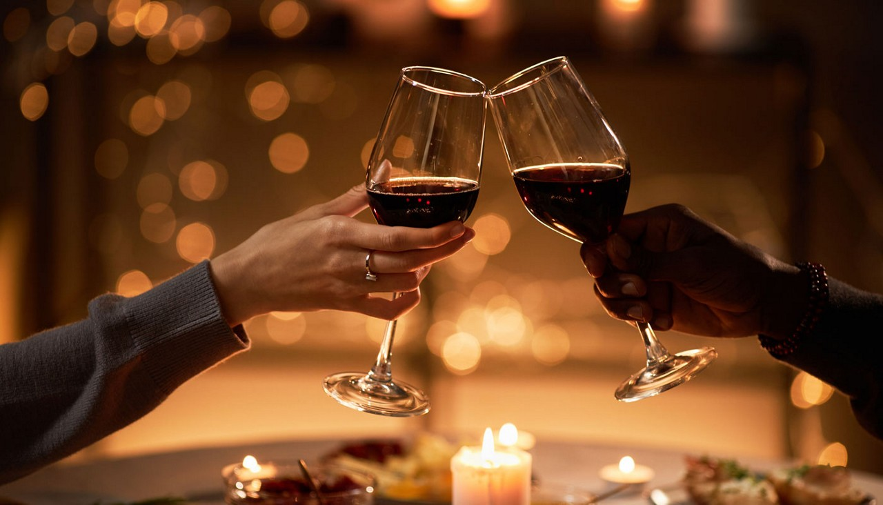 Two glasses of red wine clink over a candlelit dinner