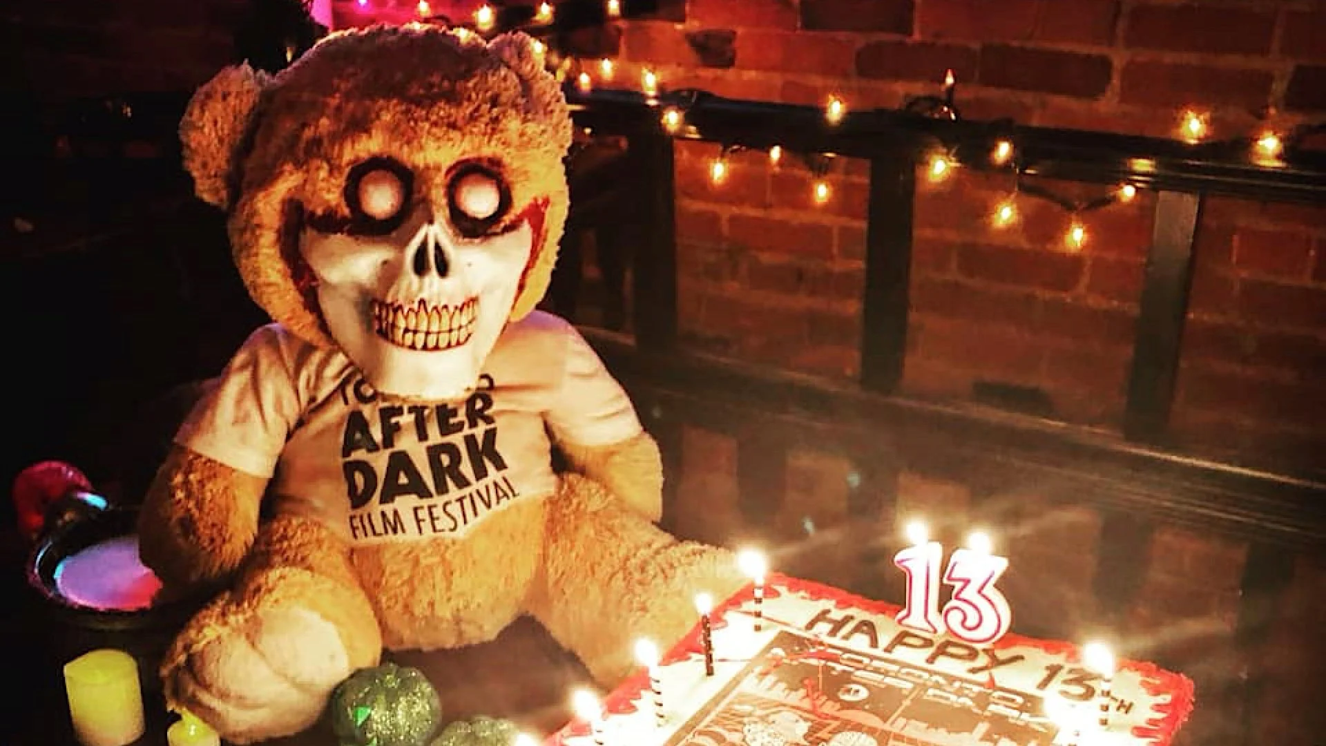 A skull-masked Toronto After Dark Film Festival mascot sits beside a cake topped with a 13 candle.